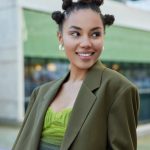 Fashionable smiling young woman with hair buns dressed in green jacket wears vivid makeup looks away happily enjoys leisure time in city poses outdoors against blurred background feels glad.