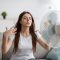 Sad young european woman suffers from unbearably too hot weather, catches cold air from fan in living room Sad young european woman suffers from unbearably too hot weather, catches cold air from fan in living room interior. Home without air conditioning, lady tired from summer heat, waves arms to cool down